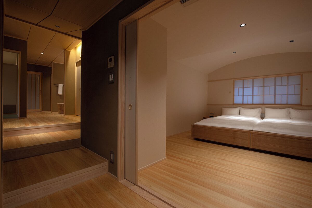 A modern bedroom with a large bed, wooden flooring, a built-in headboard, and a mirror.