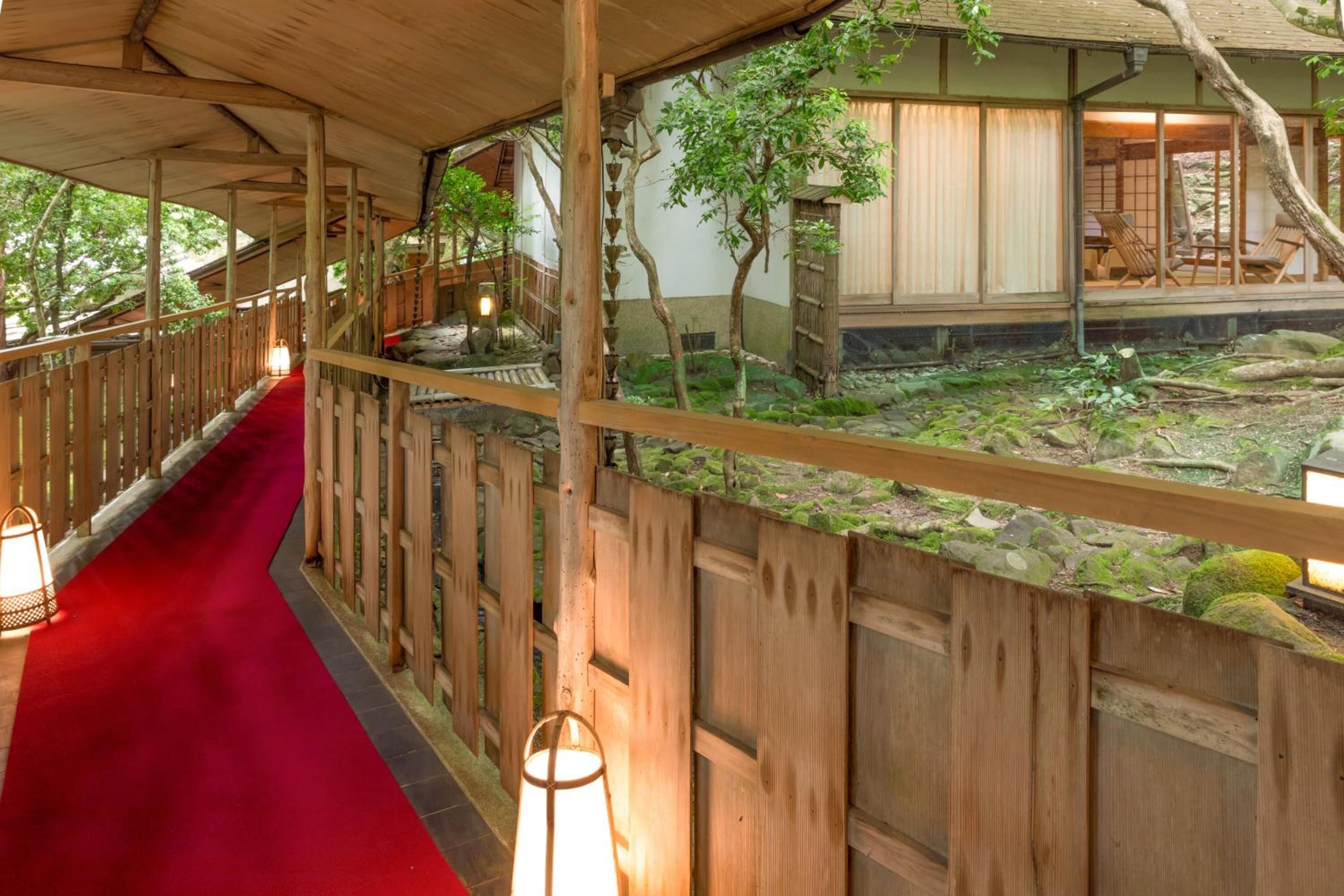 Il s'agit d'un bâtiment de style japonais traditionnel, avec une façade en bois, un chemin bordé d'une moquette rouge et une vue sur un jardin agrémenté de rochers et d'arbres.
