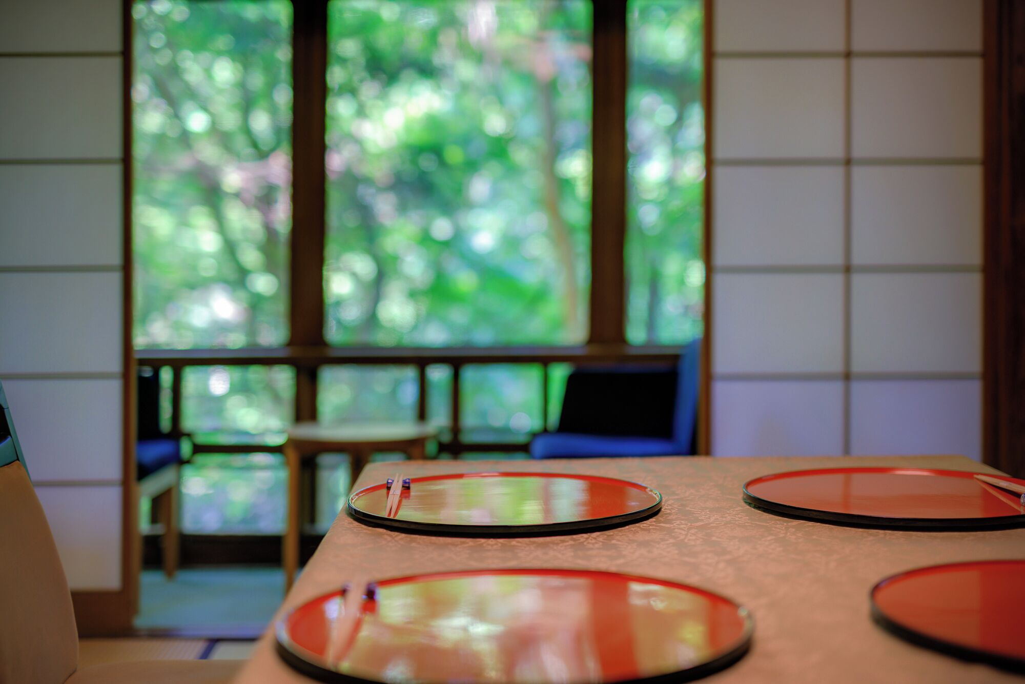 A room with a view of greenery through large windows, a wooden table with red and black plates, and a chair.