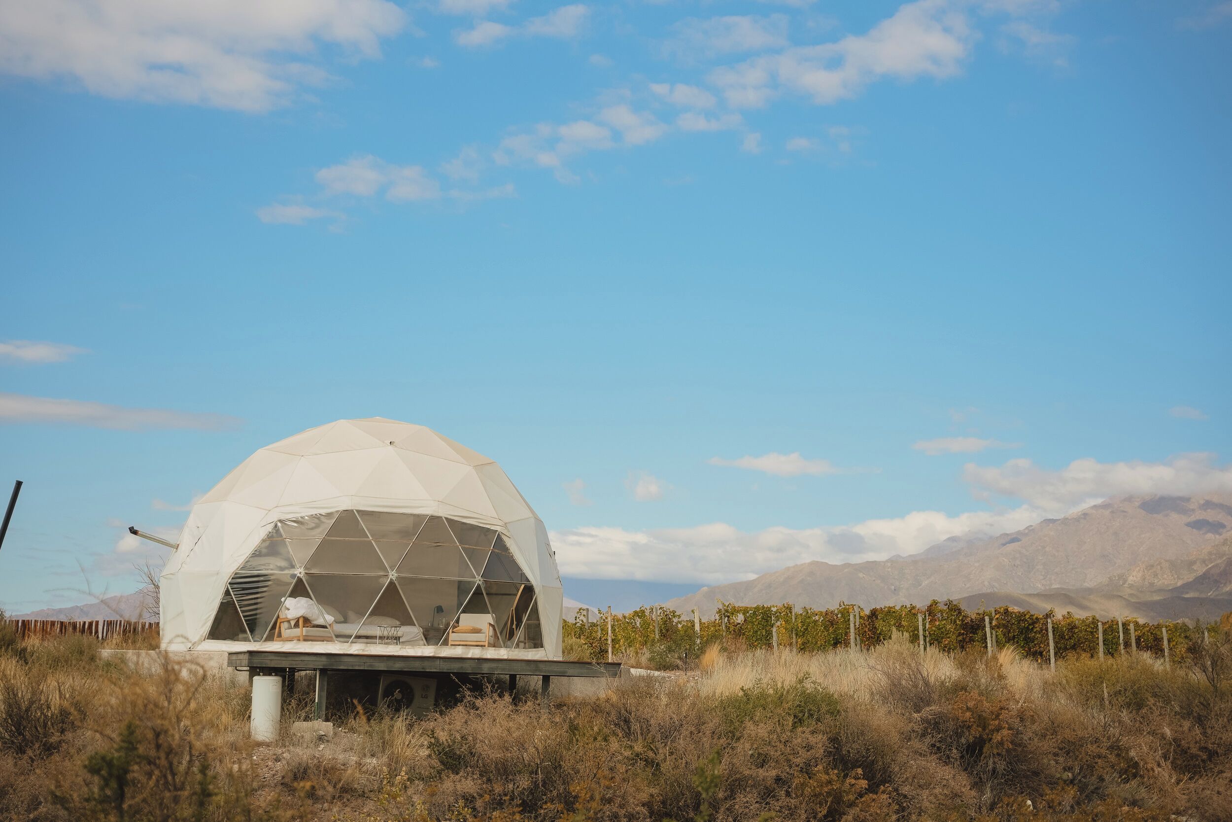 Una estructura de domo geodésico con techo plano, ubicada en un área seca y cubierta de pasto, con montañas al fondo.