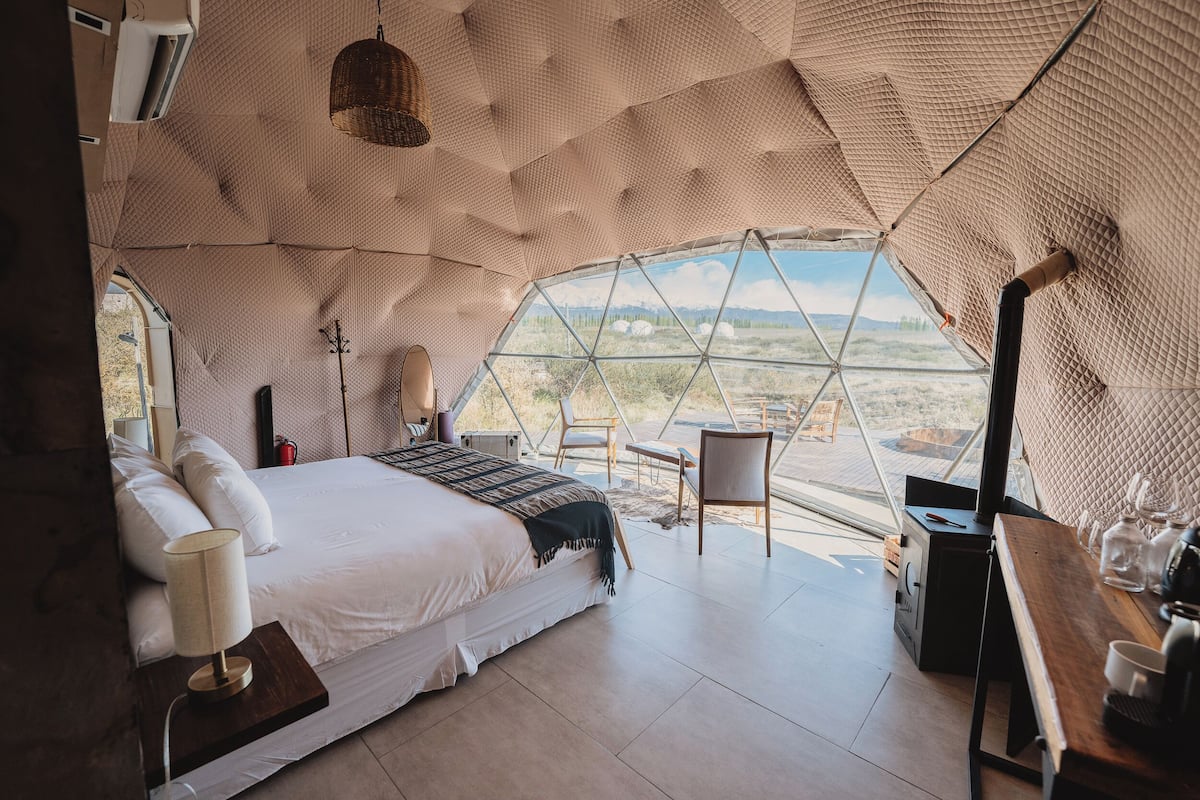 A spacious, modern bedroom with a large bed, a bedside table, a lamp, and a view of the desert through a large window.