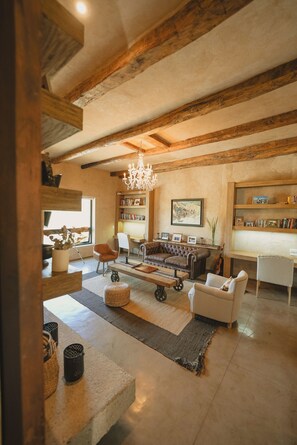 Amplia sala de estar con vigas de madera a la vista, candelabro, chimenea y estantería.