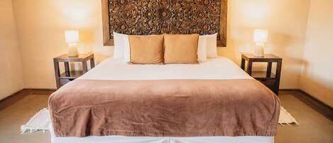 Una cama bien hecha con un cabecero marrón, dos mesitas de noche con lámparas y un panel decorativo en la pared.