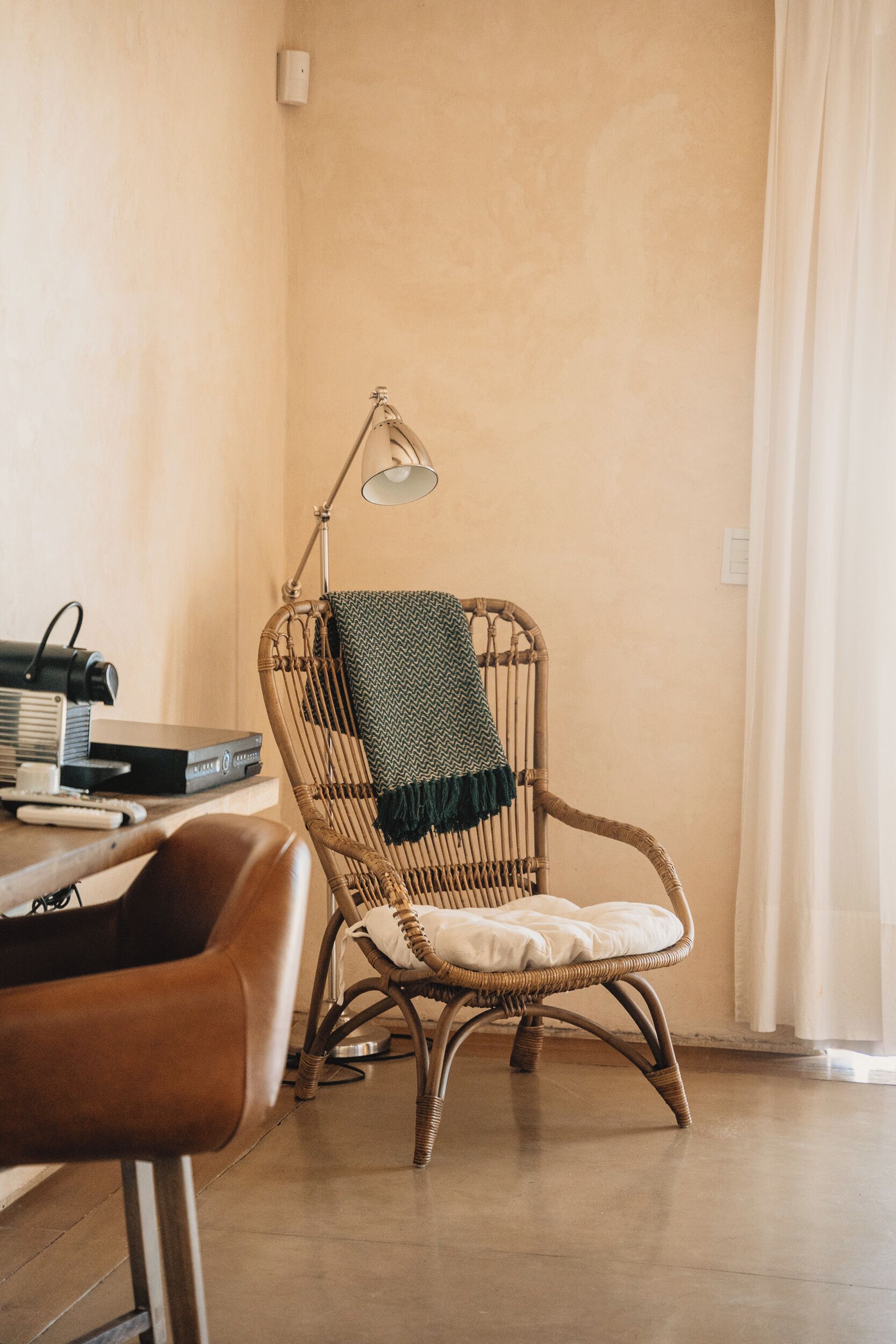Una silla de mimbre con cojín, una lámpara y una mesa con libros y un control remoto.