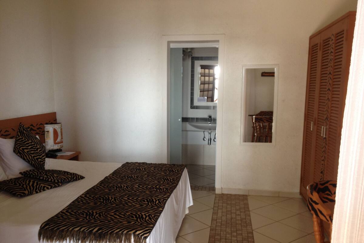 A hotel room with a bed, a nightstand, a television, a zebra-patterned blanket, a bathroom visible through an open door, and a wardrobe with a mirror.