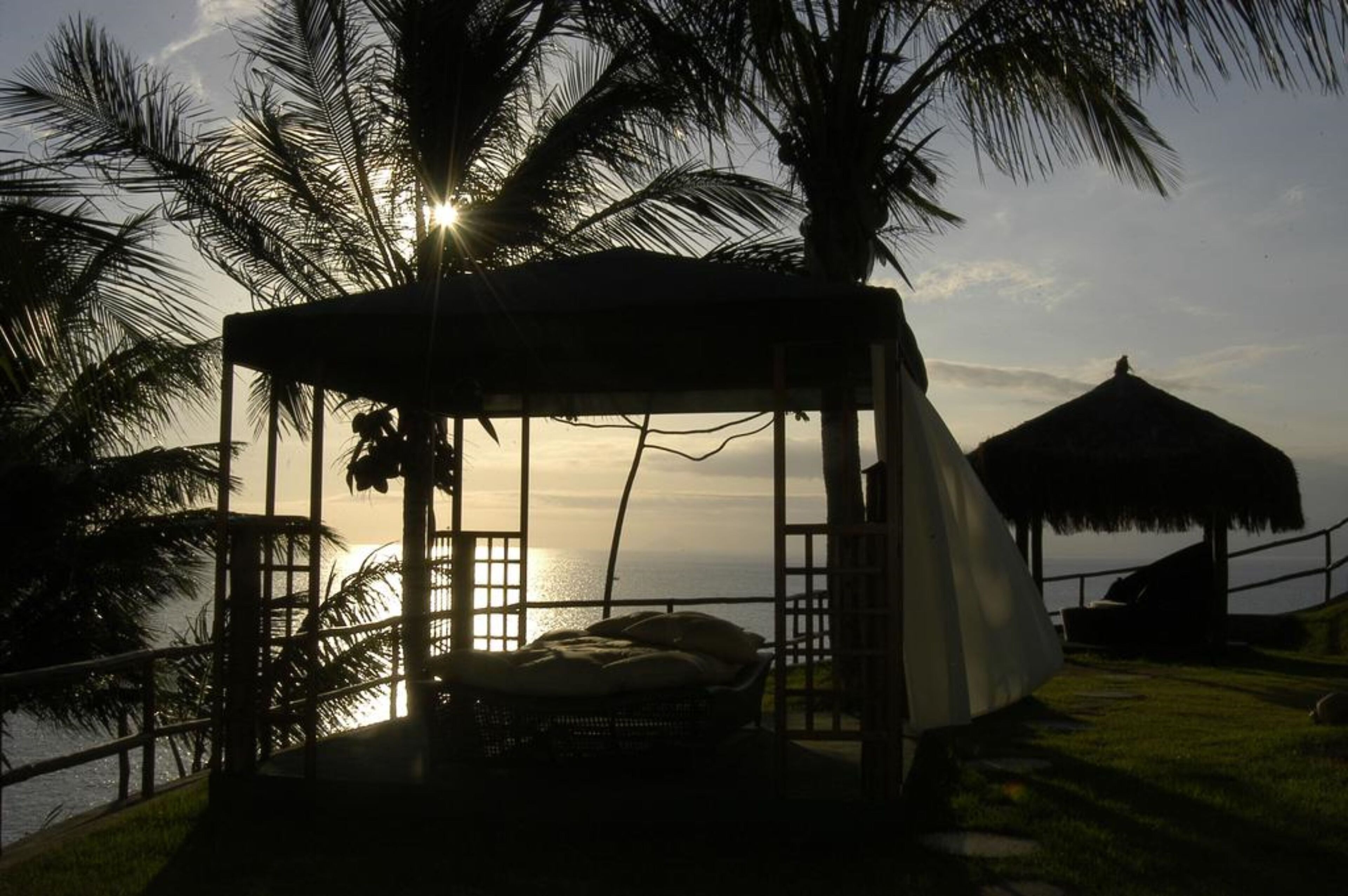 A beachside hut with a thatched roof, a bed, and a view of the ocean at sunset.
