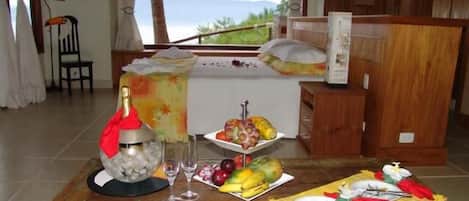 Une chambre d’hôtel avec un lit, une table à manger avec des fruits et une bouteille, et une vue sur les montagnes.