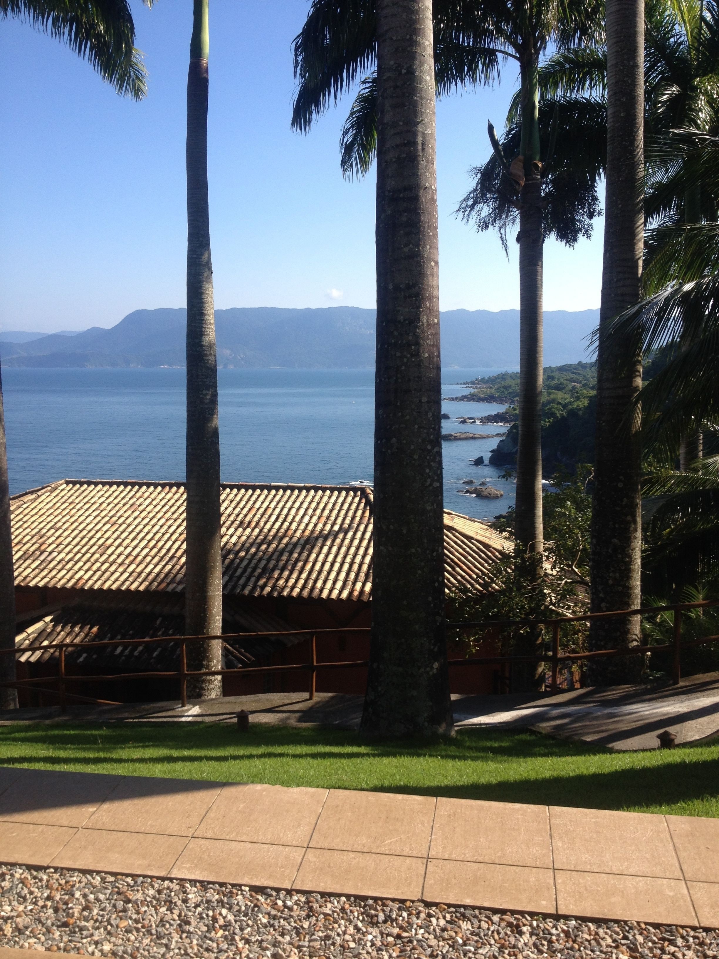 A view of a coastal area with palm trees, a tiled roof structure, and a clear view of the sea and distant mountains.