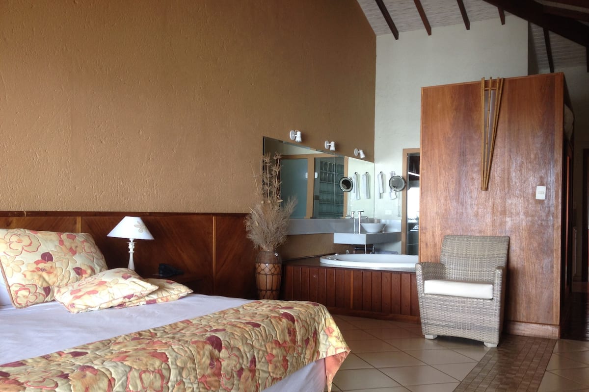 A hotel room with a large bed, a sitting area with a chair, a small kitchenette, and a wooden wardrobe.