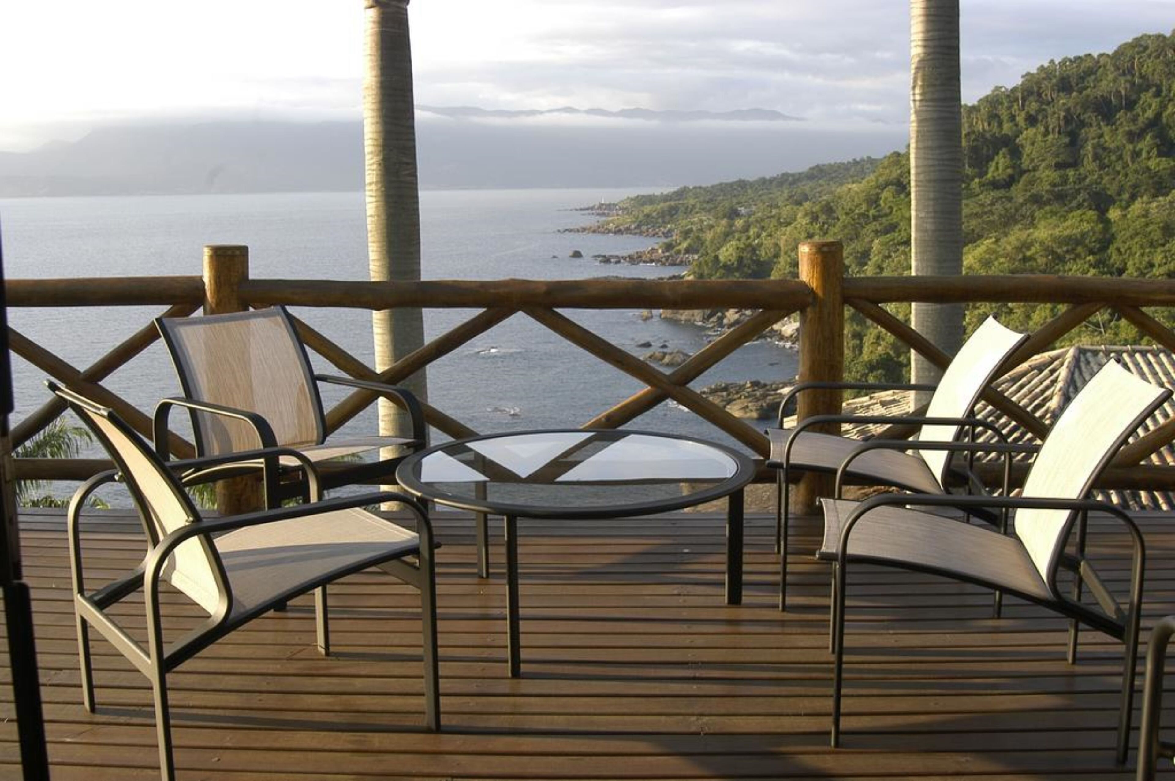 En terrasse med stole og et bord med udsigt over et kystlandskab, hvor havet og en skovklædt bakke ses.