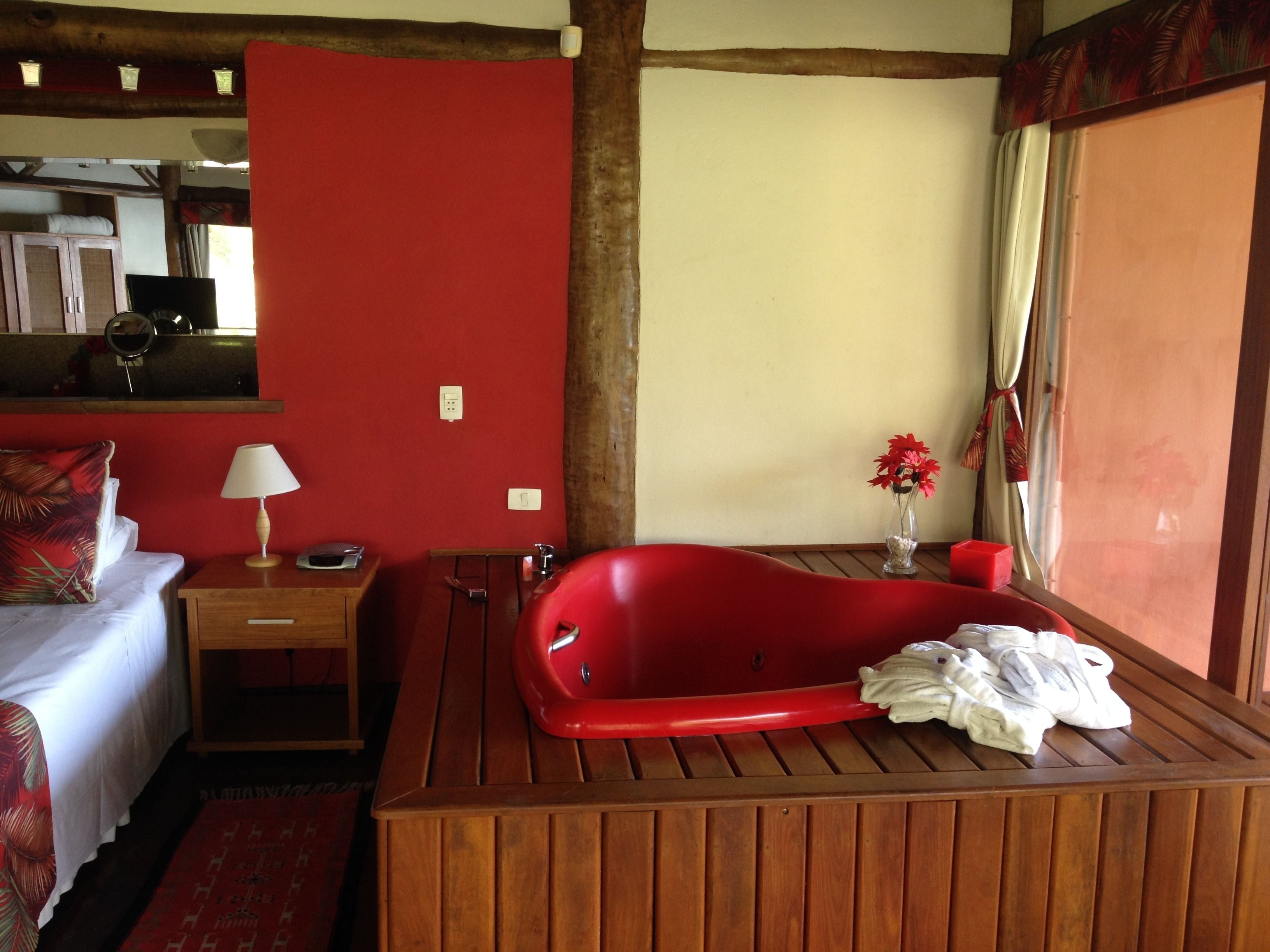 Ein Hotelzimmer mit roter Wand, einer Holzwanne, einem Bett mit rotem Kissen, einem Nachttisch mit Lampe und einem Fenster mit Vorhängen.