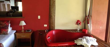Ein Hotelzimmer mit roter Wand, einer Holzwanne, einem Bett mit rotem Kissen, einem Nachttisch mit Lampe und einem Fenster mit Vorhängen.