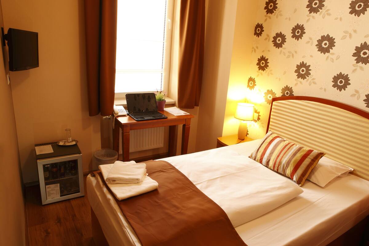 Une chambre d’hôtel comprenant un lit, un bureau avec un ordinateur portable, un petit réfrigérateur, une fenêtre avec des rideaux, un mur recouvert d’un papier peint à motifs floraux et une lampe de chevet.