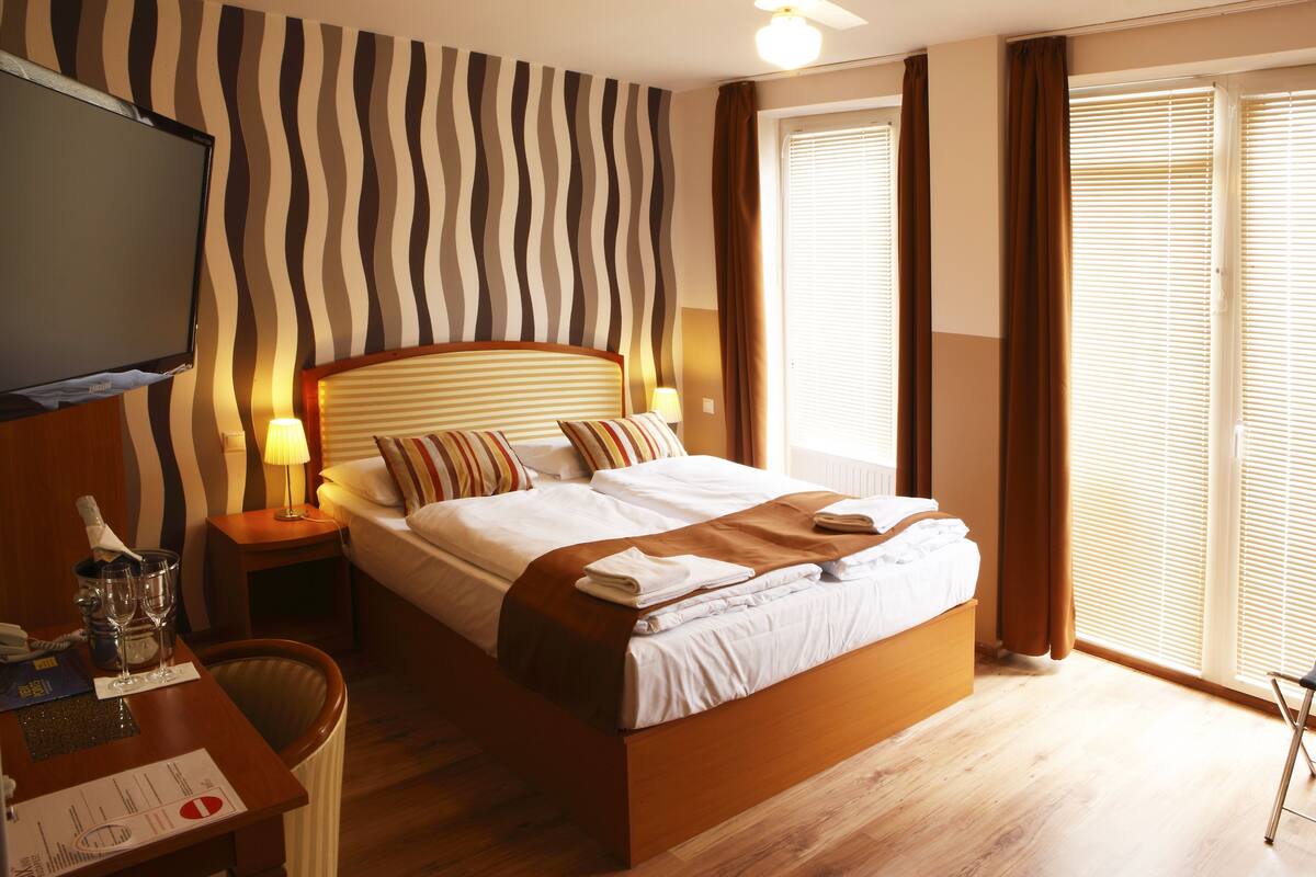 A hotel room with a bed, bedside tables, a TV, and a window with curtains.
