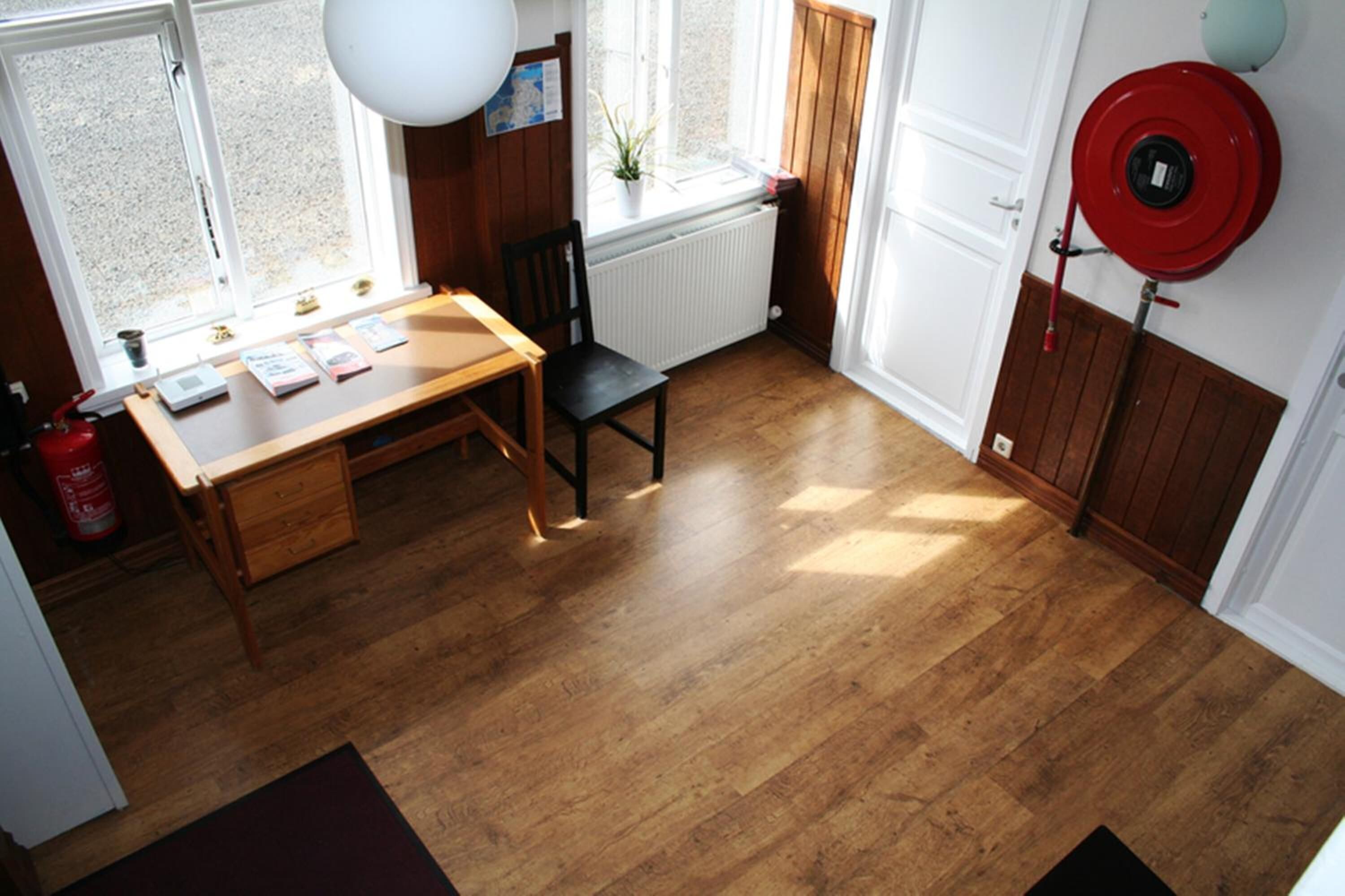 A room with a wooden floor, a desk with papers, a chair, a radiator, and a red fire extinguisher.