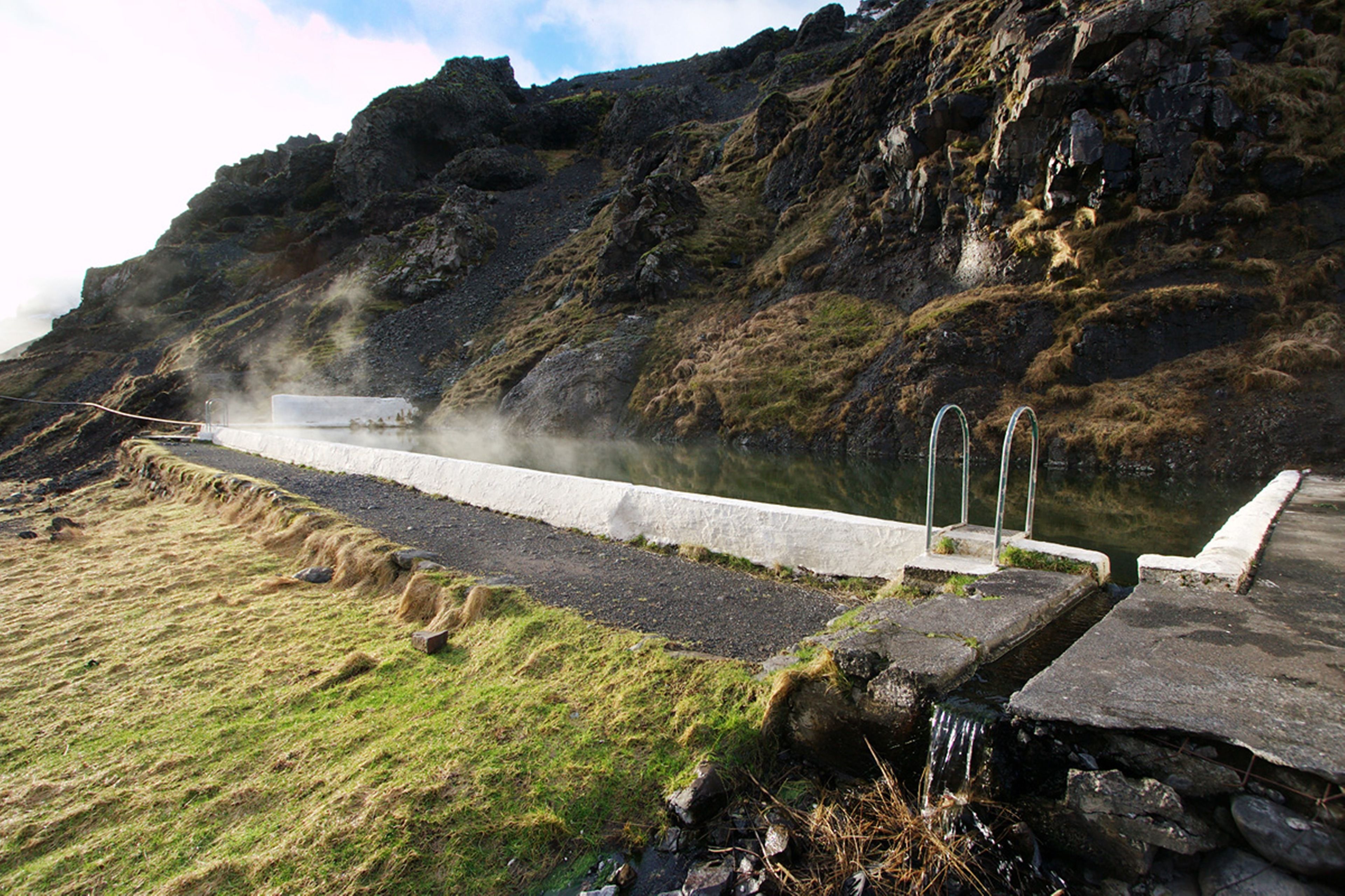 A natural hot spring with a concrete pathway and metal railings, surrounded by rocky terrain and grass.