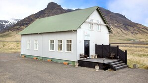 Un pequeño edificio de un piso con techo verde y paredes blancas, que cuenta con un porche con banco y mesa, con montañas al fondo.