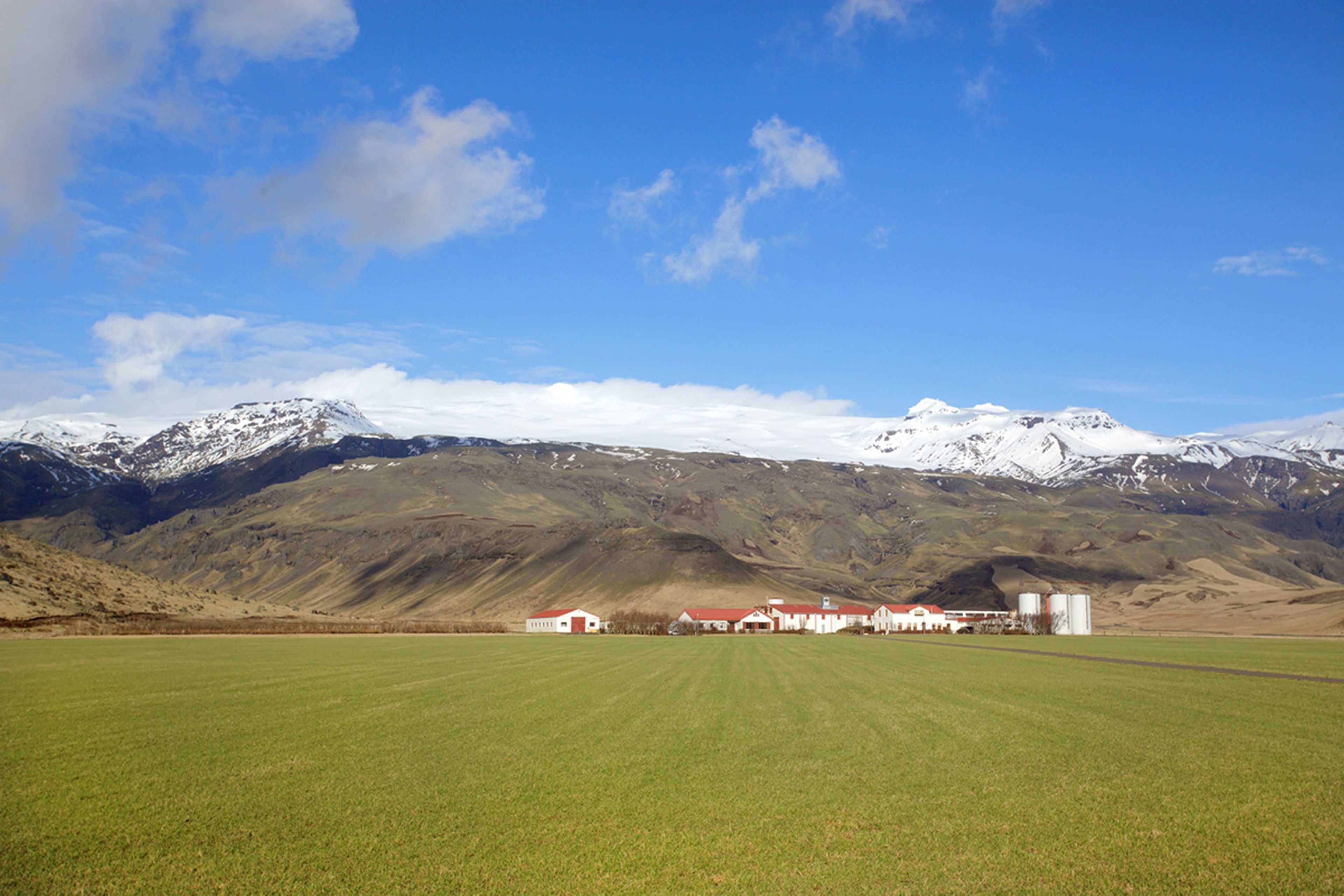 A rural landscape with a green field, a row of white buildings with red roofs, and snow-capped mountains in the background.