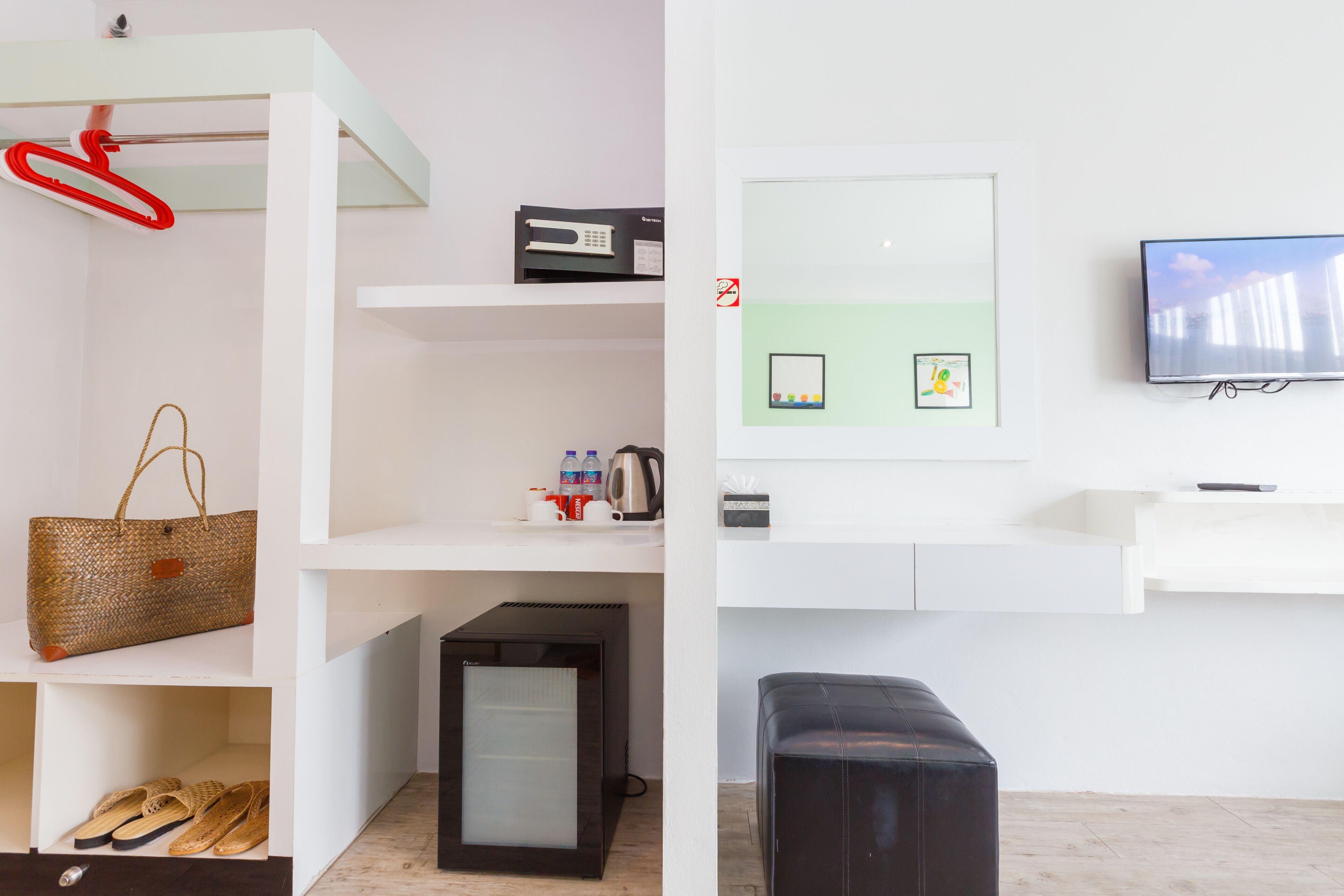 Une chambre moderne avec un mur blanc, un téléviseur à écran plat, un petit réfrigérateur noir, un panier en osier, une paire de sandales et un pouf noir.