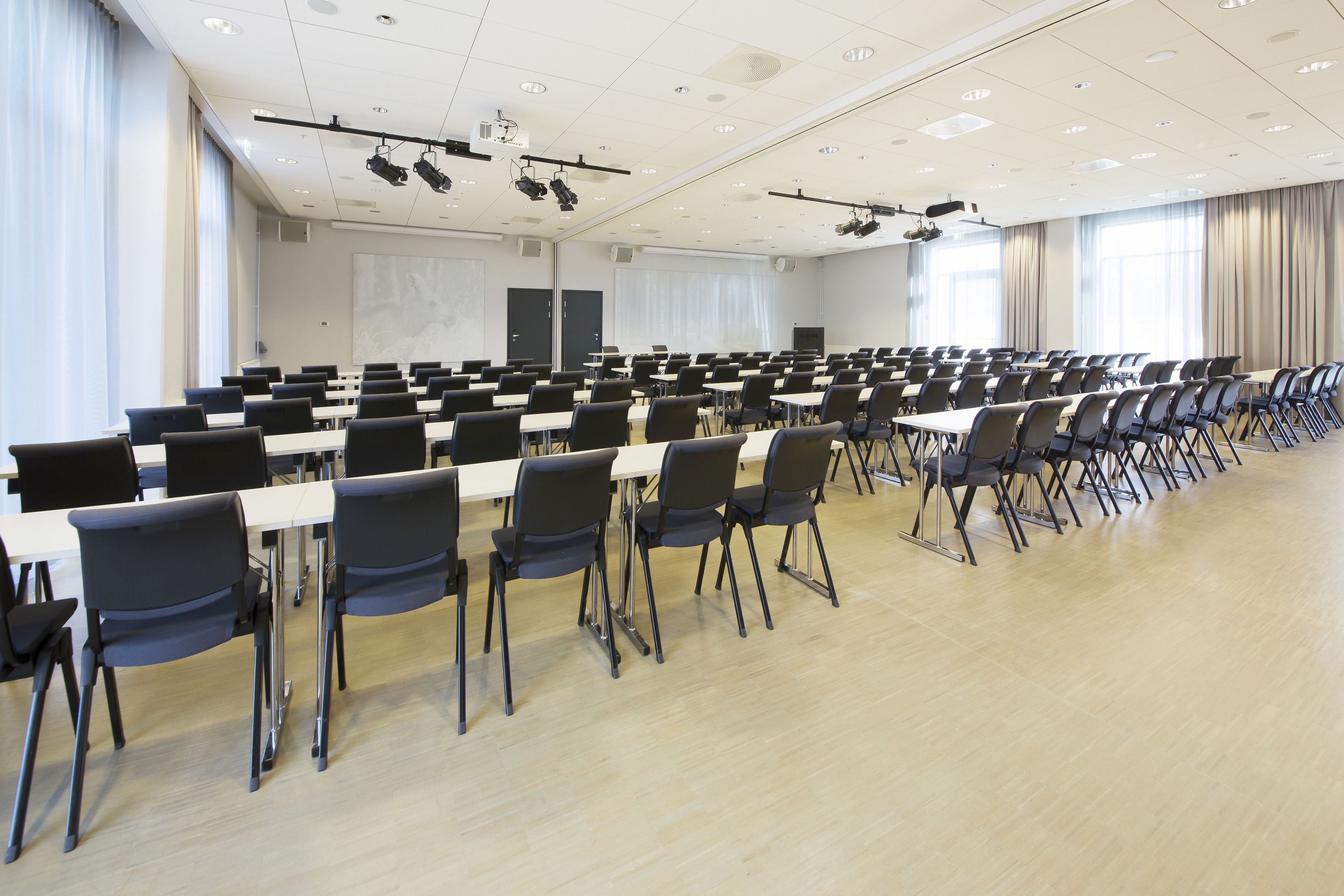 Ein großer Konferenzraum mit Reihen von Tischen und Stühlen, ausgestattet mit Deckenprojektoren und Lautsprechern.