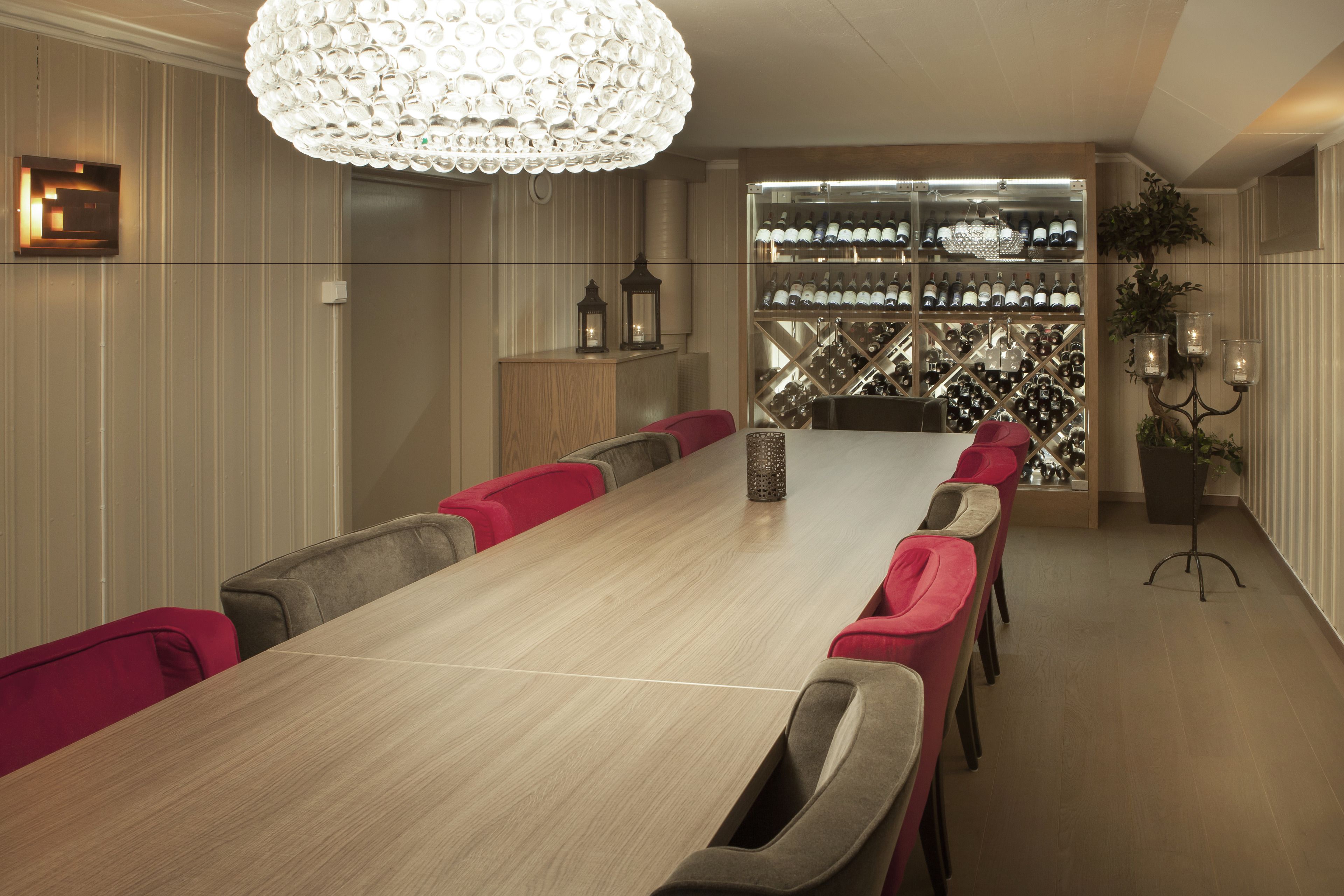 A conference room with a long table, red and gray chairs, a chandelier, and a wine rack in the background.