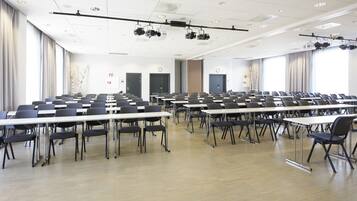 Un salón de conferencias con filas de mesas y sillas, proyectores instalados en el techo y ventanas amplias con cortinas.