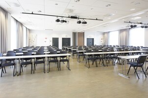 Un salón de conferencias con filas de mesas y sillas, proyectores instalados en el techo y ventanas amplias con cortinas.