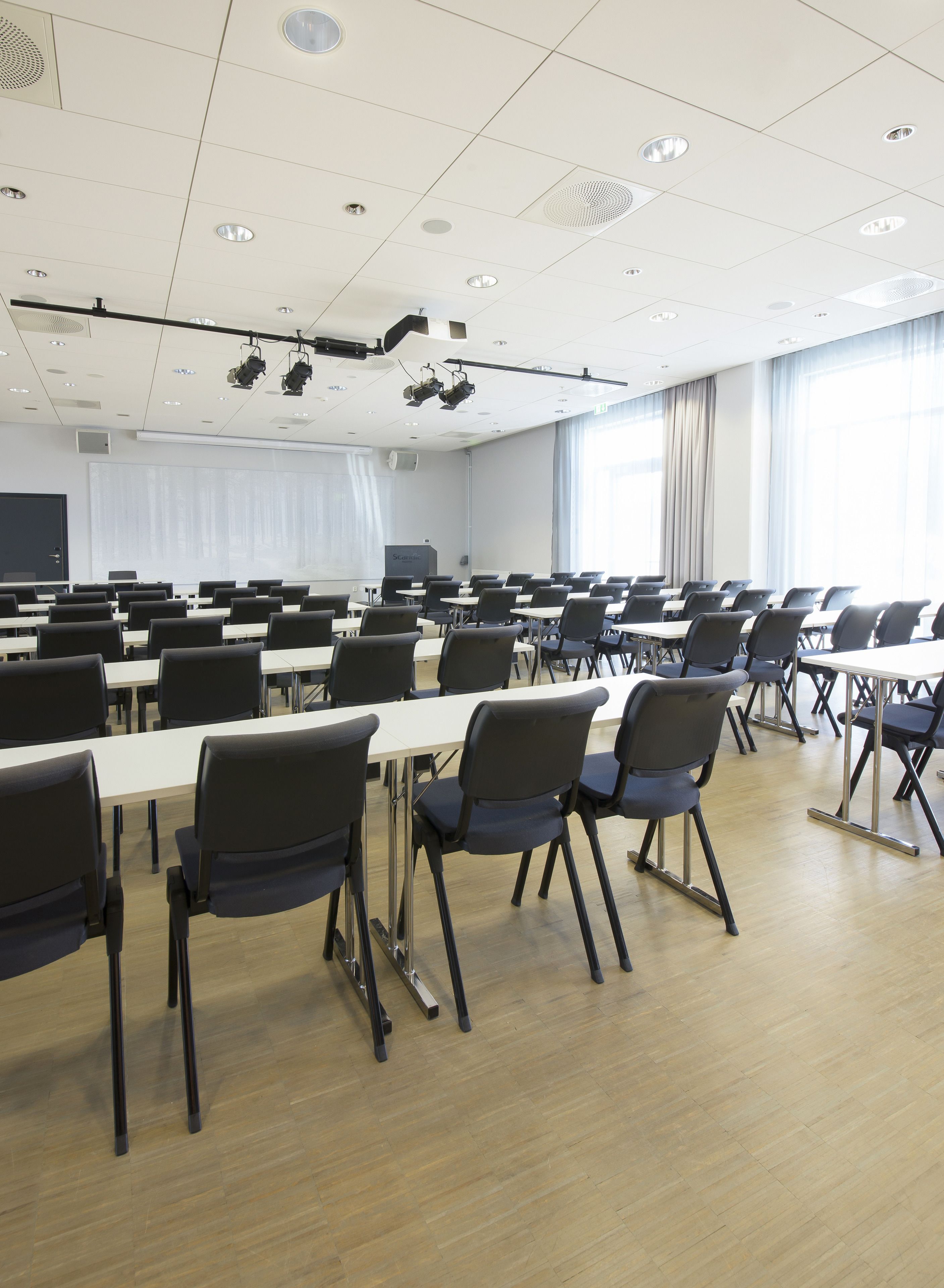 Un salón de conferencias con mesas y sillas dispuestas en filas, un proyector y una pantalla.