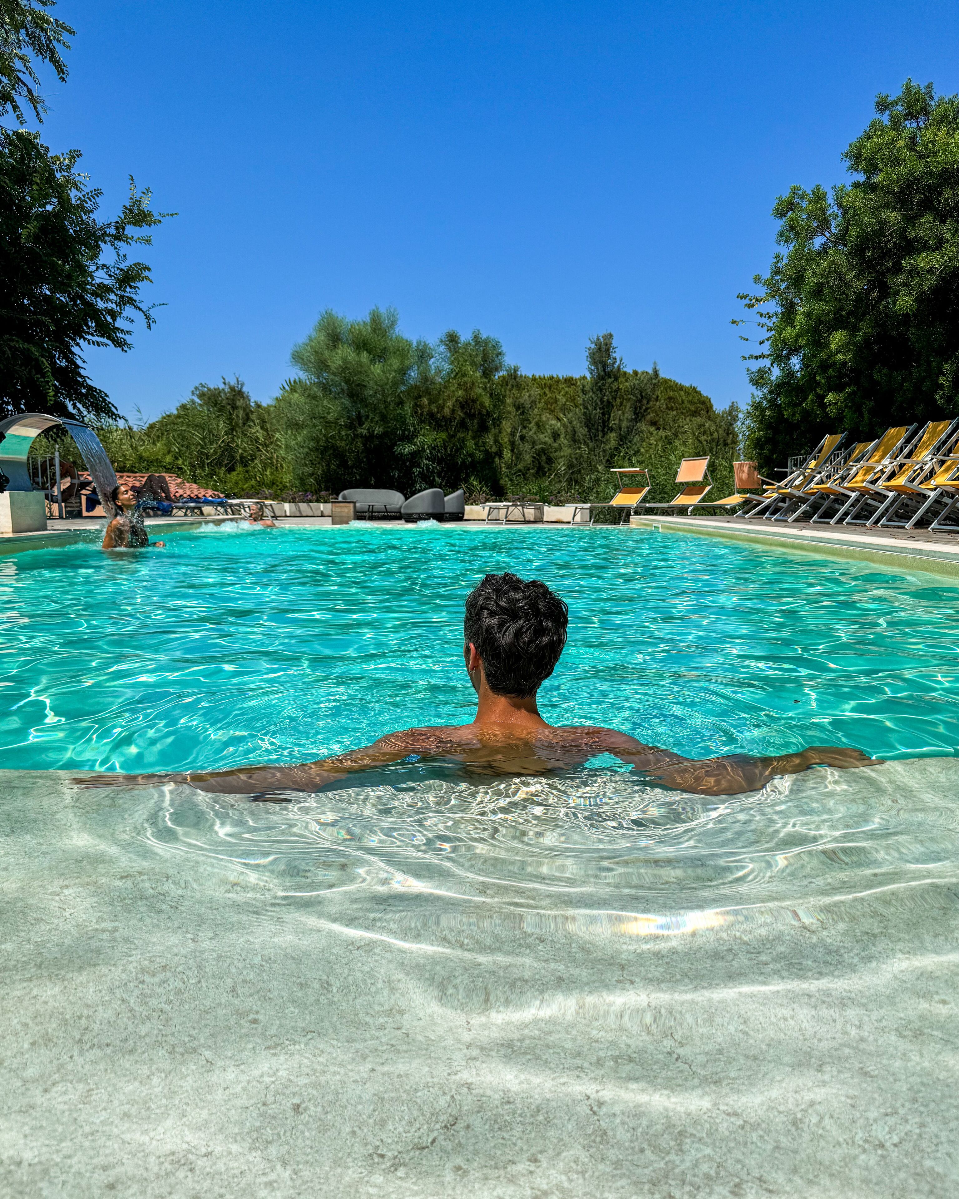 Un uomo in piscina, con acqua cristallina, circondato da sdraio e da un'area a bordo piscina.
