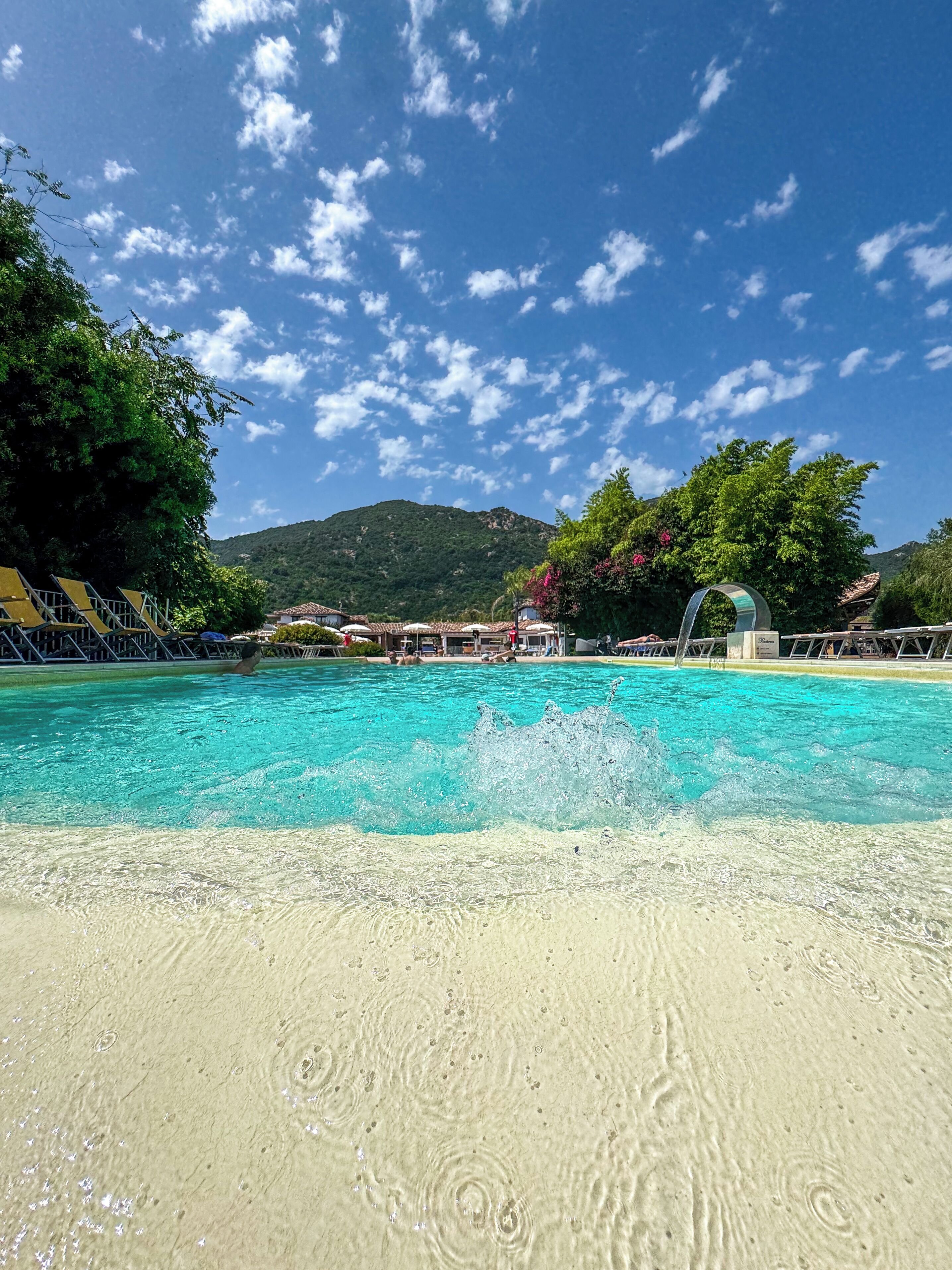 A clear blue pool with a sandy bottom, surrounded by greenery and a mountain in the background.