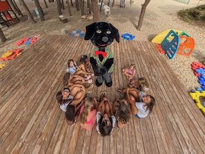 Eine Gruppe Kinder sitzt im Kreis auf einer Holzterrasse, im Zentrum befindet sich eine Maskotte.
