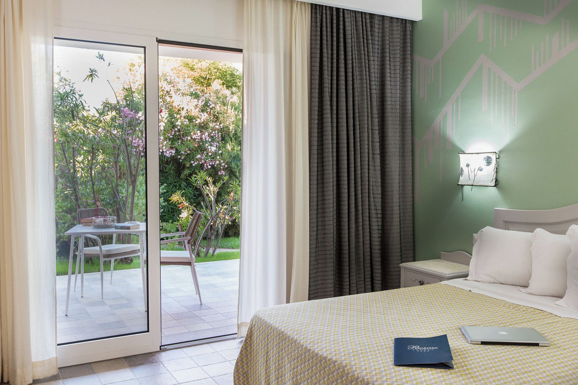 Habitación de hotel con una cama, una computadora portátil y un libro sobre la cama. Hay una ventana con cortinas y vistas a zonas verdes.