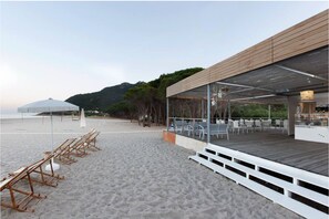 A beachside restaurant with outdoor seating, a covered area, and a view of the beach and hills in the background. - Perdepera Resort (Cardedu)