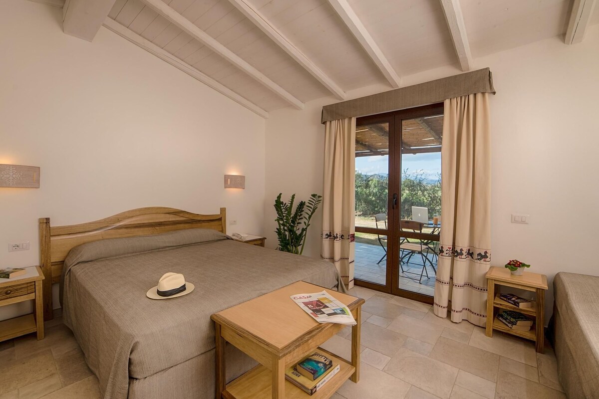 Een slaapkamer met een houten bed, een klein tafeltje met een tijdschrift, een hoed en uitzicht op het landschap door een schuifdeur.