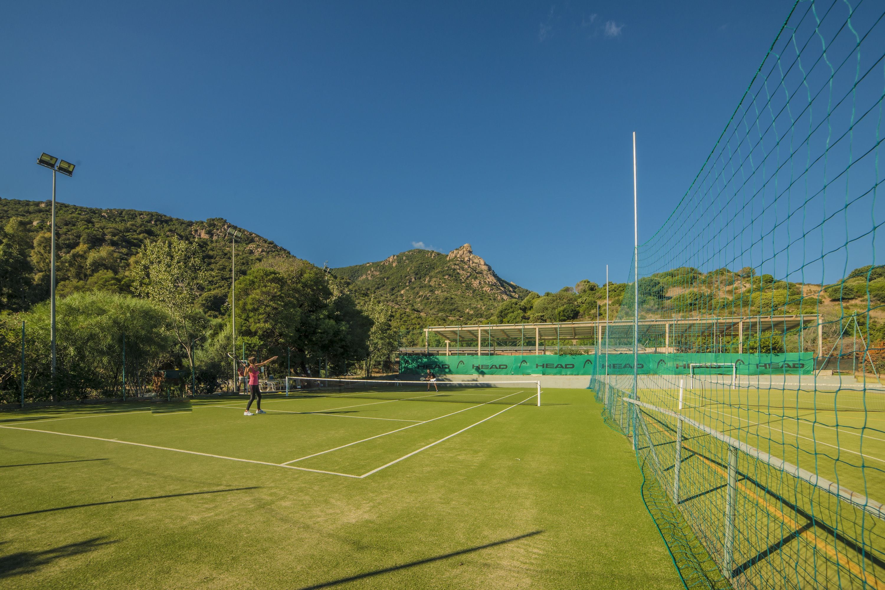 A tennis court with a net, surrounded by a fence, and a mountainous backdrop.
