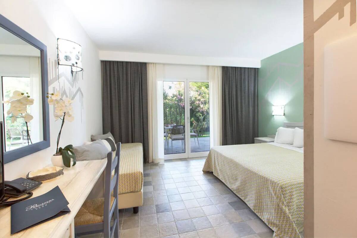 Ein Hotelzimmer mit Schreibtisch, zwei Betten, einem großen Fenster mit Vorhängen, einem Balkon und einer Wandleuchte.
