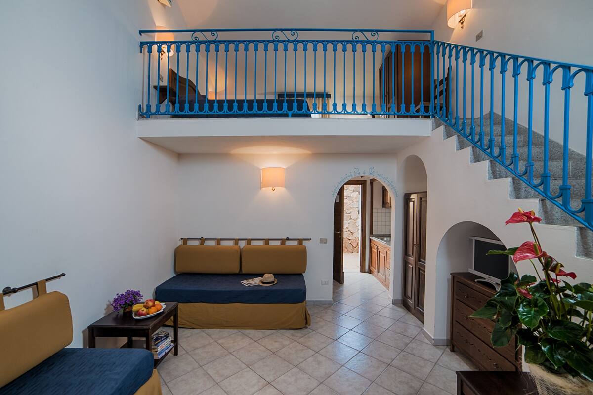 Ein modernes Wohnzimmer mit einer Couch, einem kleinen Tisch mit Obstschale und einem TV-Schrank. Eine Treppe mit blauen Geländern führt auf eine erhöhte Ebene.