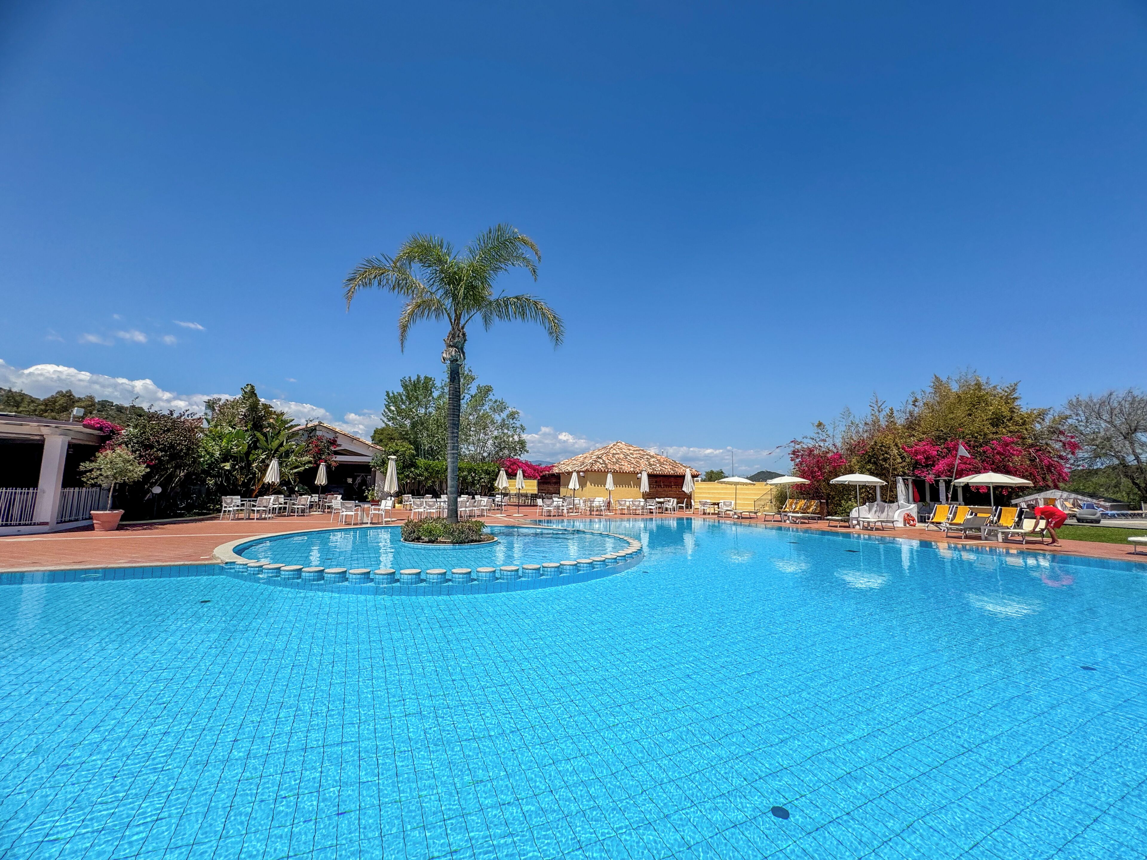 Ein Swimmingpool mit einer Palme, Liegestühlen und einem Gebäude mit einem Ziegeldach.