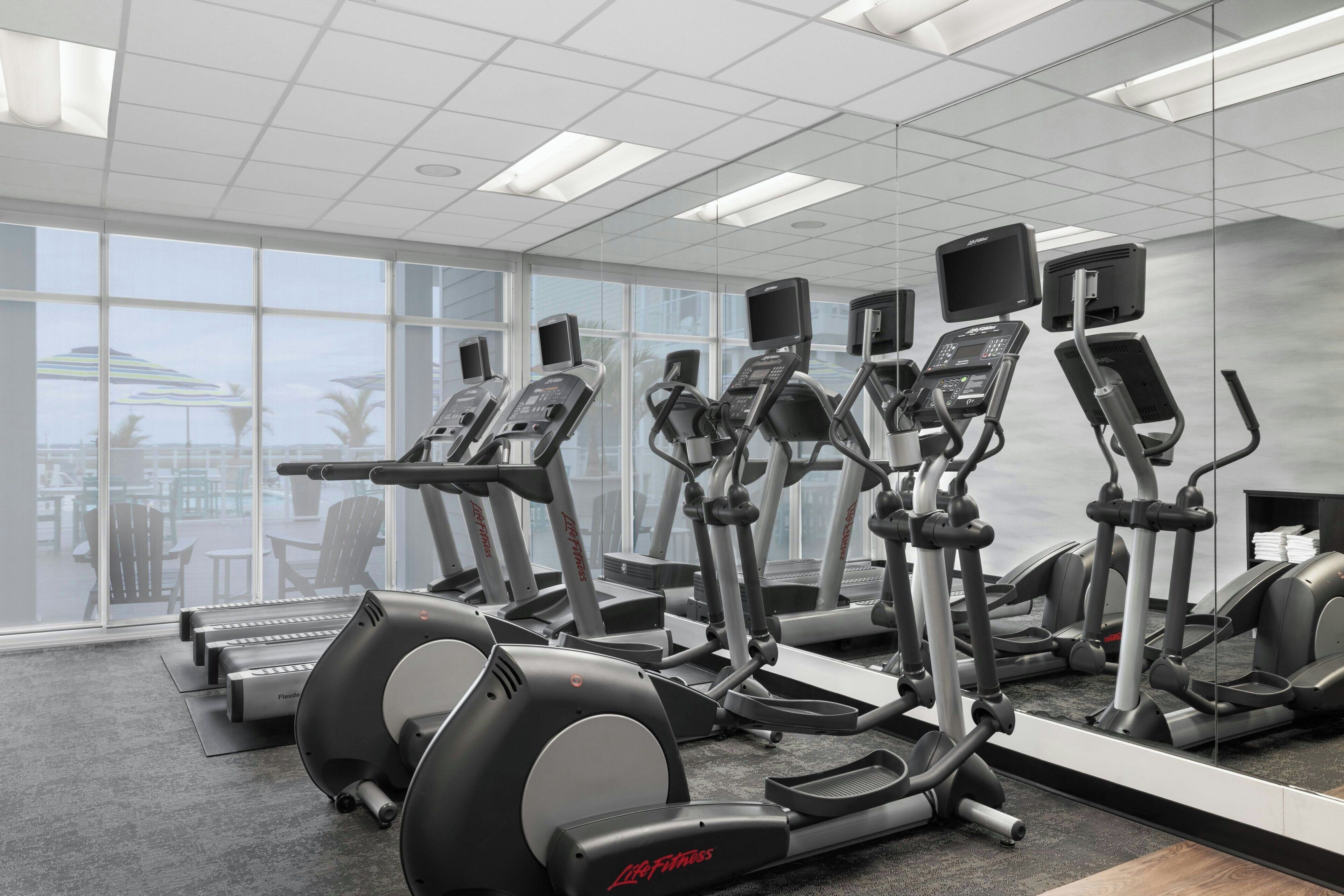A gym with various cardio machines, large windows, and a view of outdoor seating.