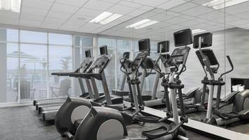 A gym with various cardio machines, large windows, and a view of outdoor seating.