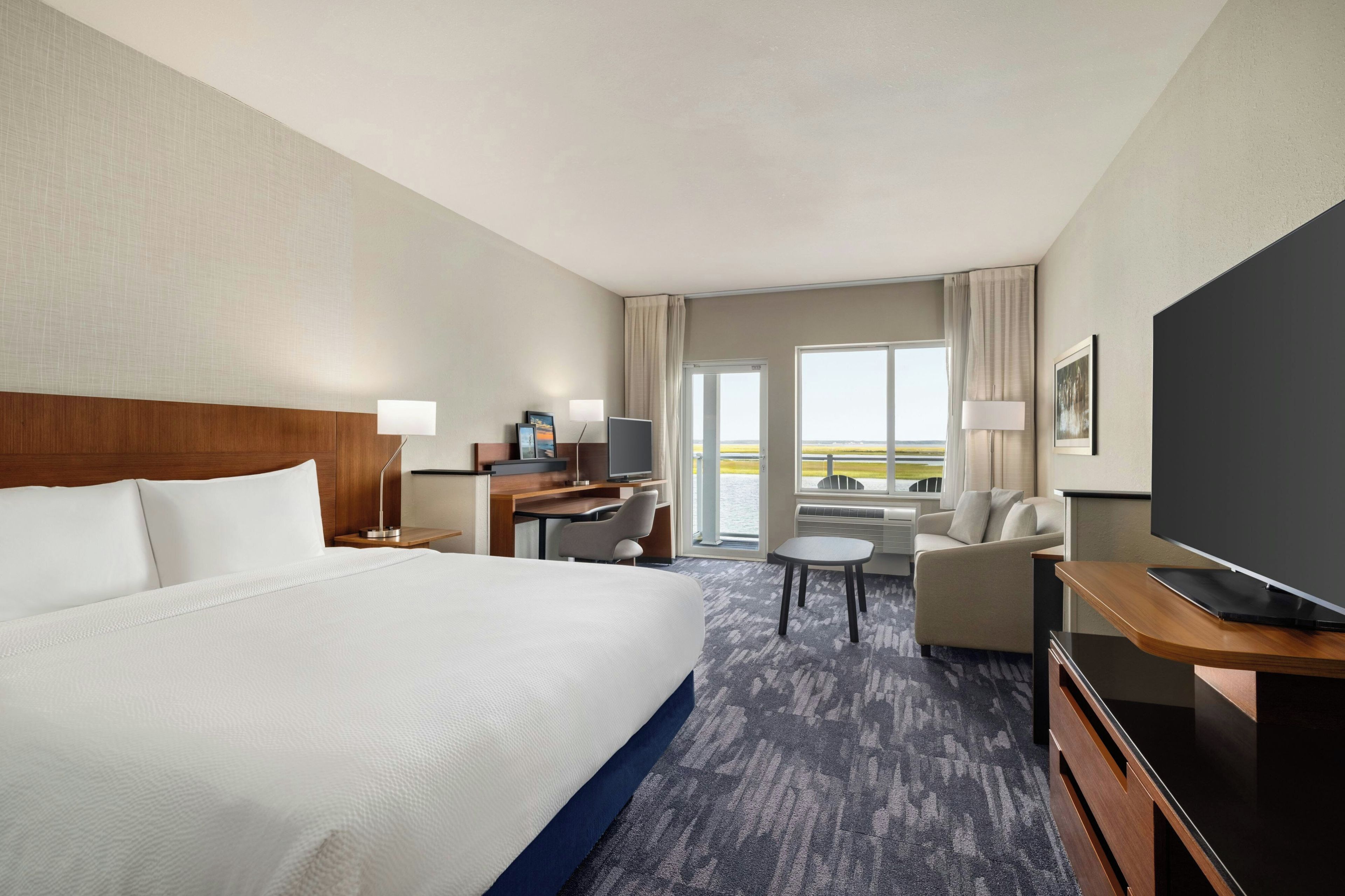 A hotel room with a large bed, a desk with a computer, a TV, a chair, and a view of the outdoors.
