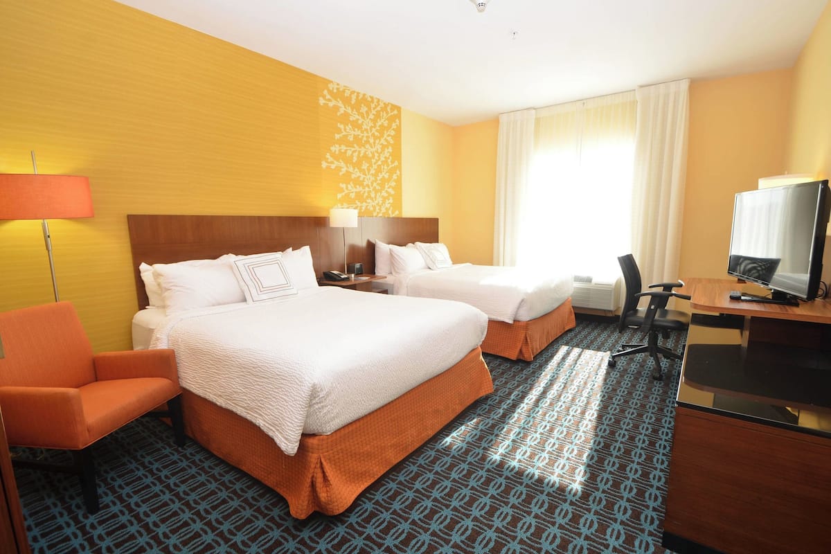 A hotel room with two beds, a desk, a chair, a TV, and a lamp.