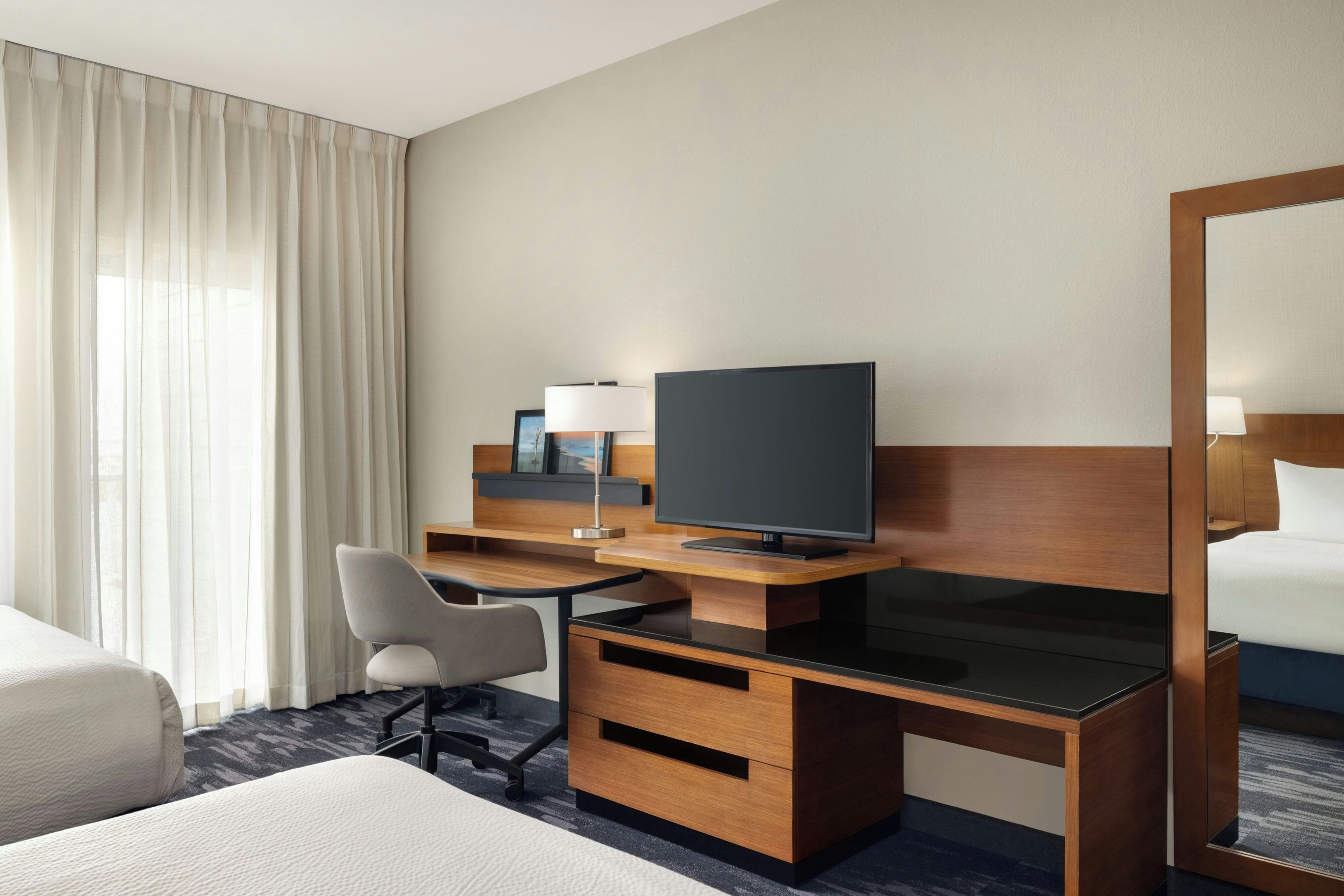 Habitación de hotel con cama, escritorio con computadora, una silla, televisor y un espejo grande.
