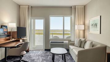 A hotel room with a desk, chair, sofa, and a view of the outdoors through large windows.