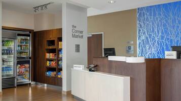 A hotel reception area with a self-service kiosk, a refrigerator stocked with beverages, and a sign for 'the Corner Market' indicating nearby amenities.
