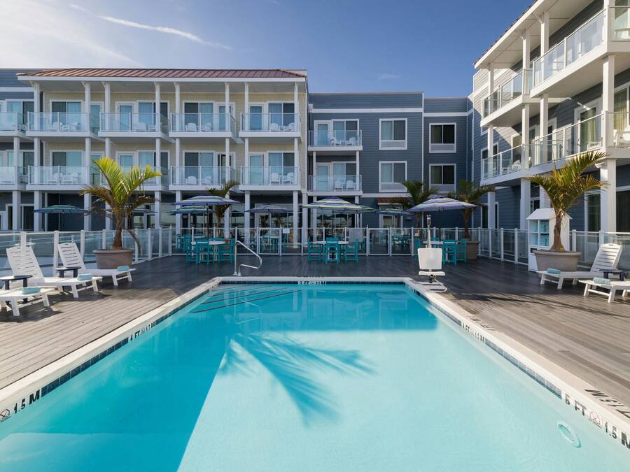 A pool area with lounge chairs and umbrellas, a clear blue pool, and a multi-story building with balconies.