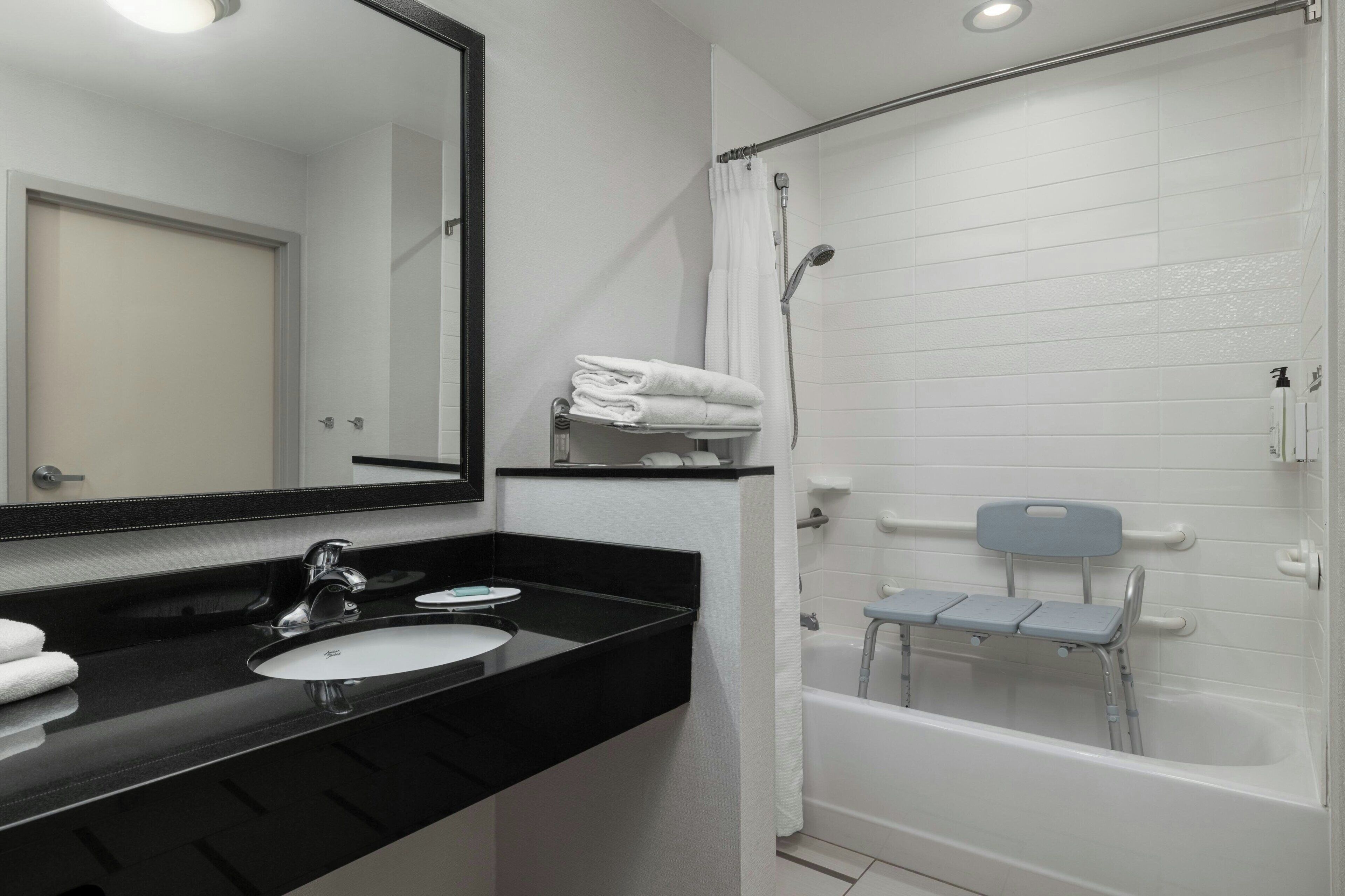Un baño con lavamanos de encimera negra, un espejo, ducha con asiento y toallero.
