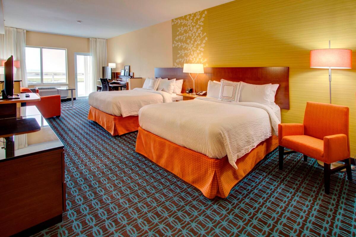 A hotel room with two beds, a desk, a chair, and a lamp.