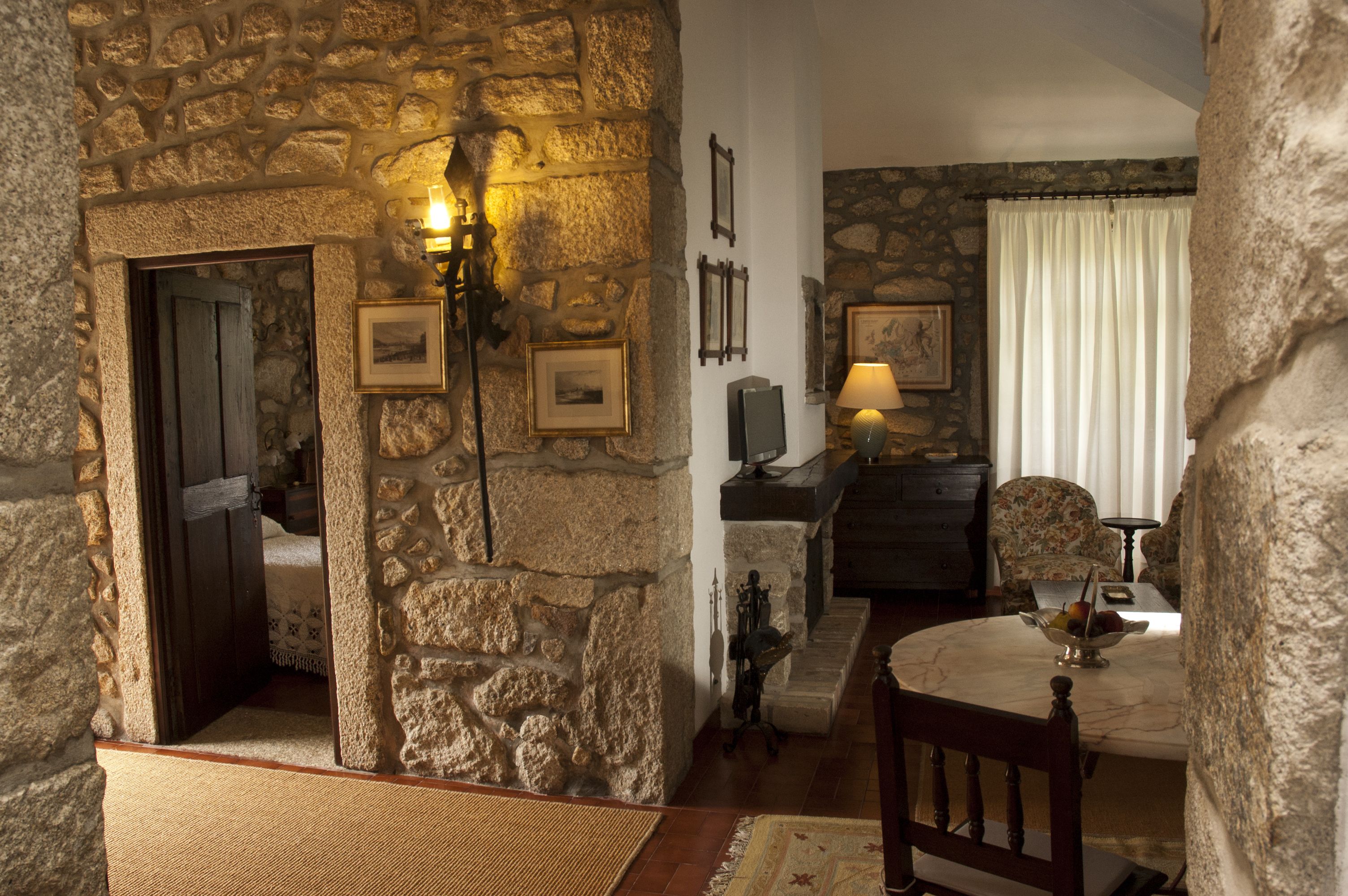 A stone-walled room with a wooden door, a table with a bowl of fruit, and a lamp.