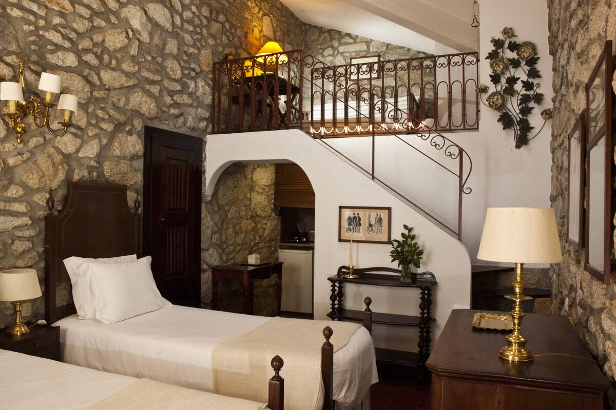 Un dormitorio con pared de piedra, una cama de madera, una escalera y una mesita de noche con lámpara.